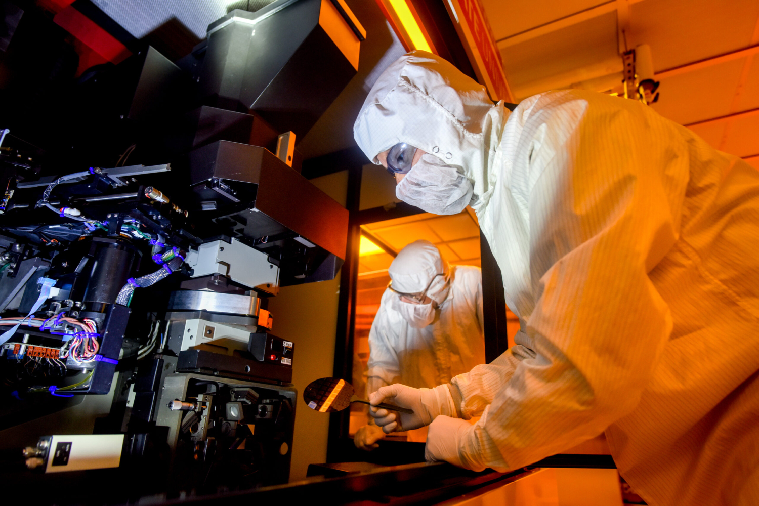 Two lab technicians dressed in head-to-toe white protective suits work in a nanofabrication facility.