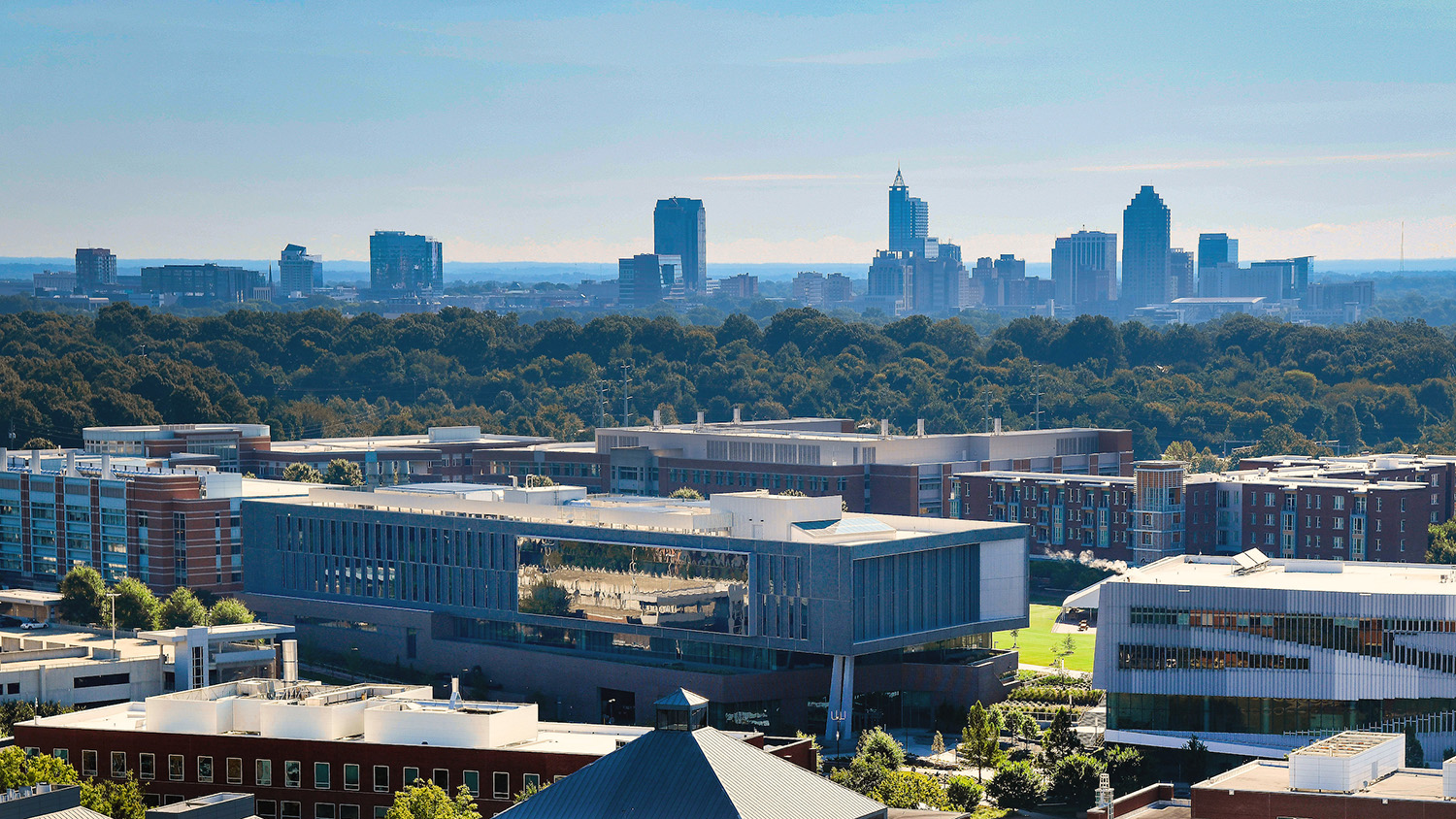 An aerial photo shows Centennial Campus buildings in the foreground and the Raleigh skyline in the background.