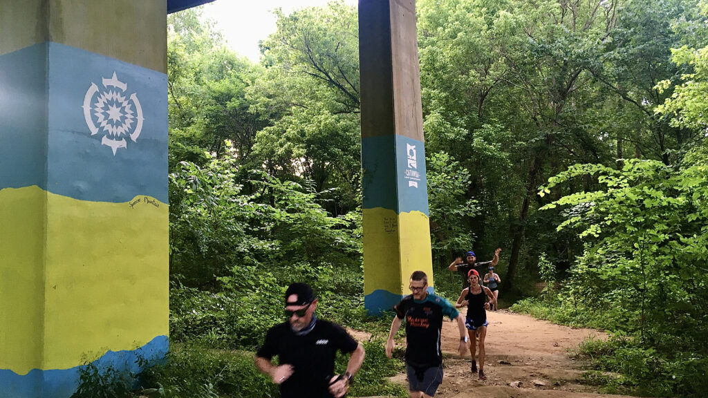 A group runners pass under bridge while traveling along a dirt trail.