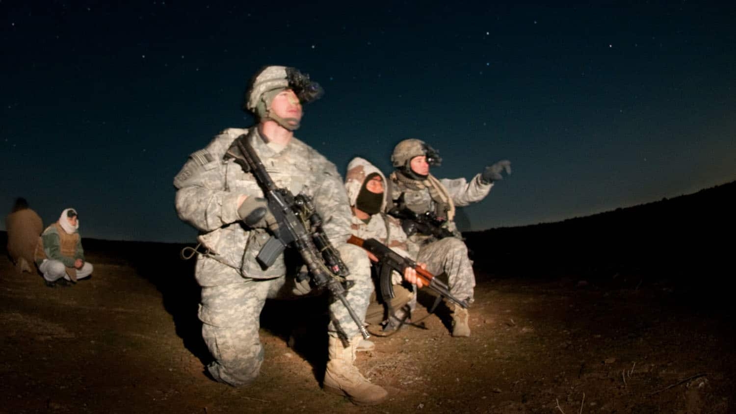 at night in the desert, a soldier with a rifle kneels in the foreground, while two other soldiers converse behind him