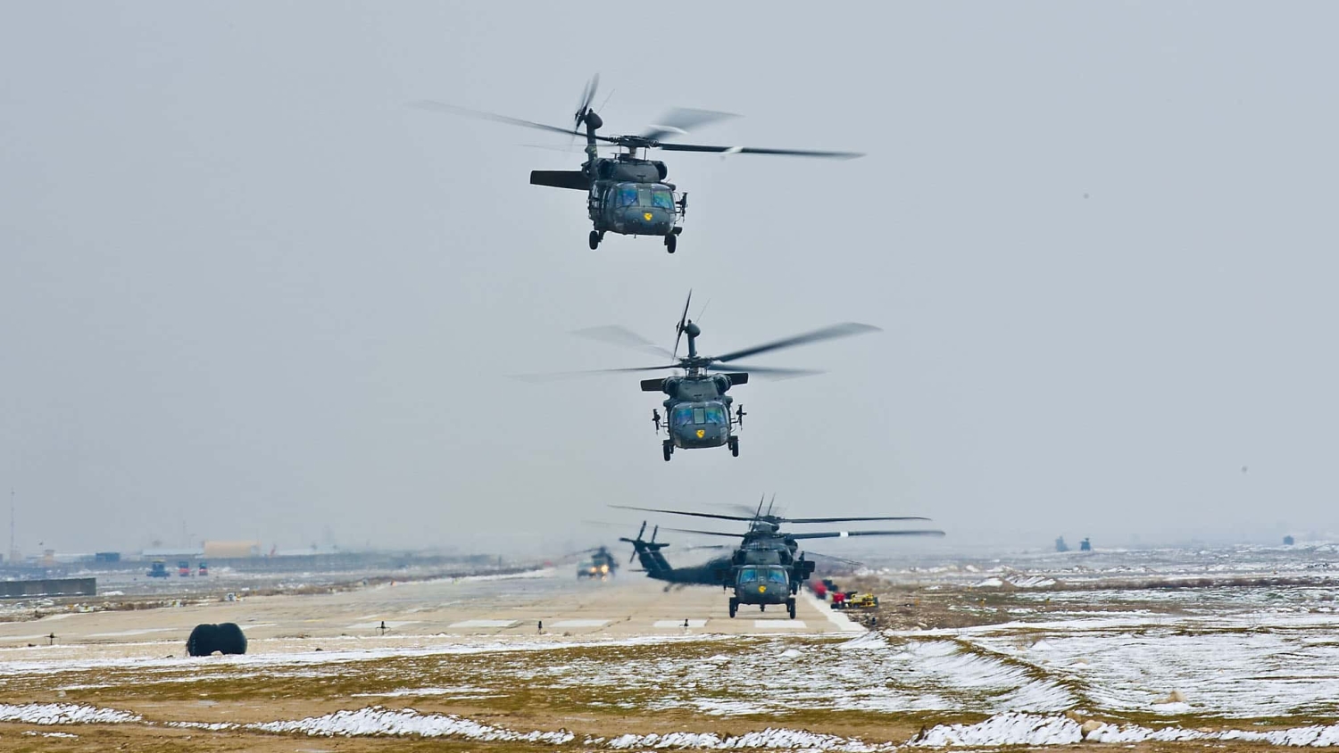 four military helicopters are taking off very close to each other on a snowy airfield
