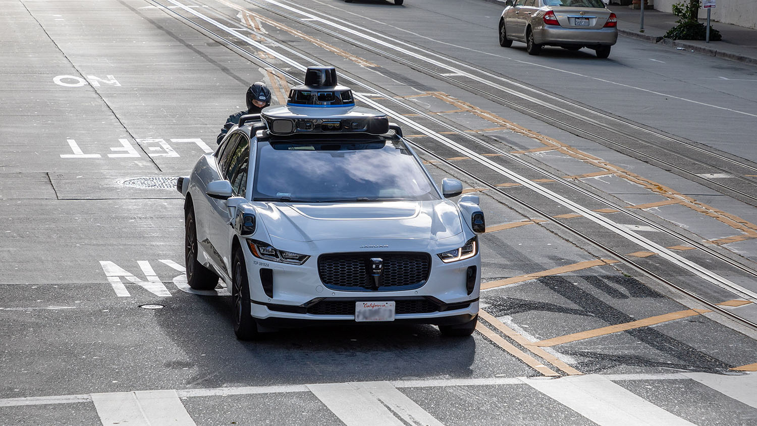 a white sedan with electronic equipment mounted on the roof and corners of the vehicle sits at an intersection. The vehicle is marketed as a "driverless car."